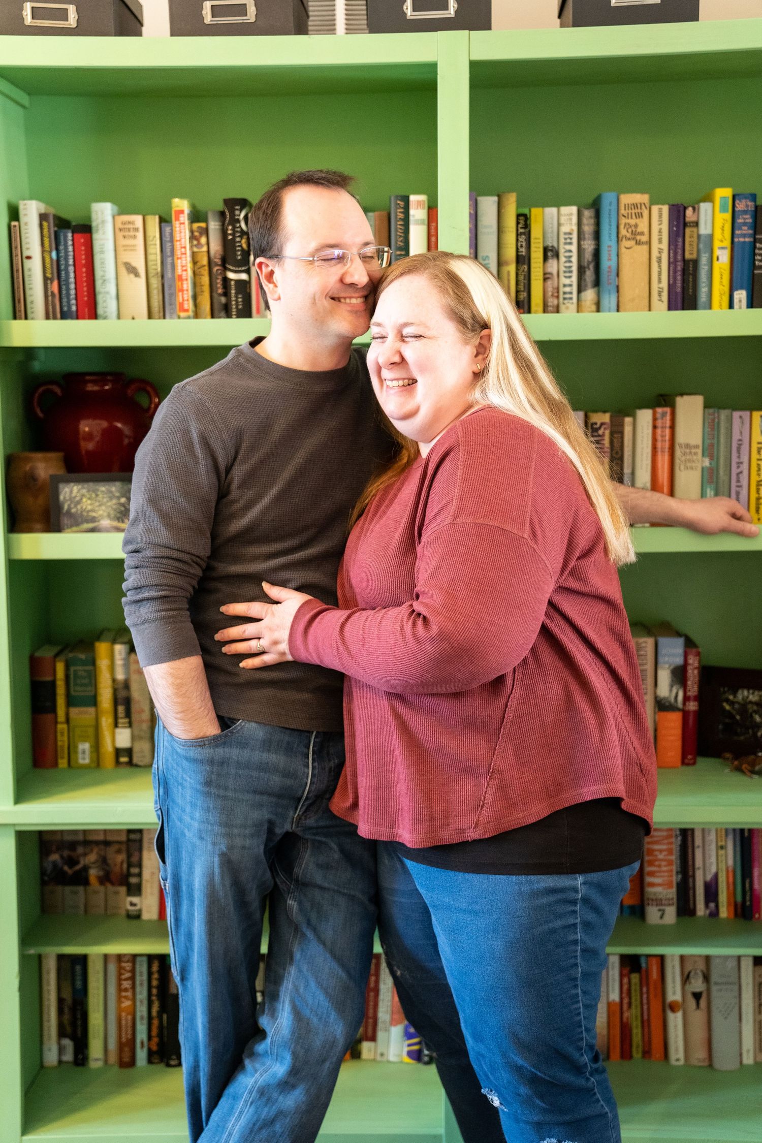 Family portrait of man wearing a brown sweater and women wearing a red sweater in front of a green bookcase laughing and smiling.