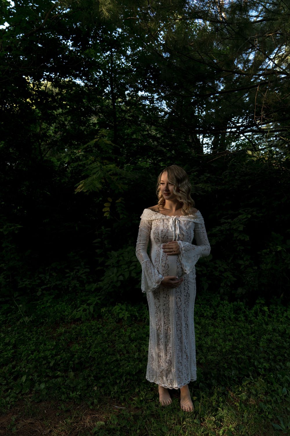A person in a white lace dress poses against lush green foliage.