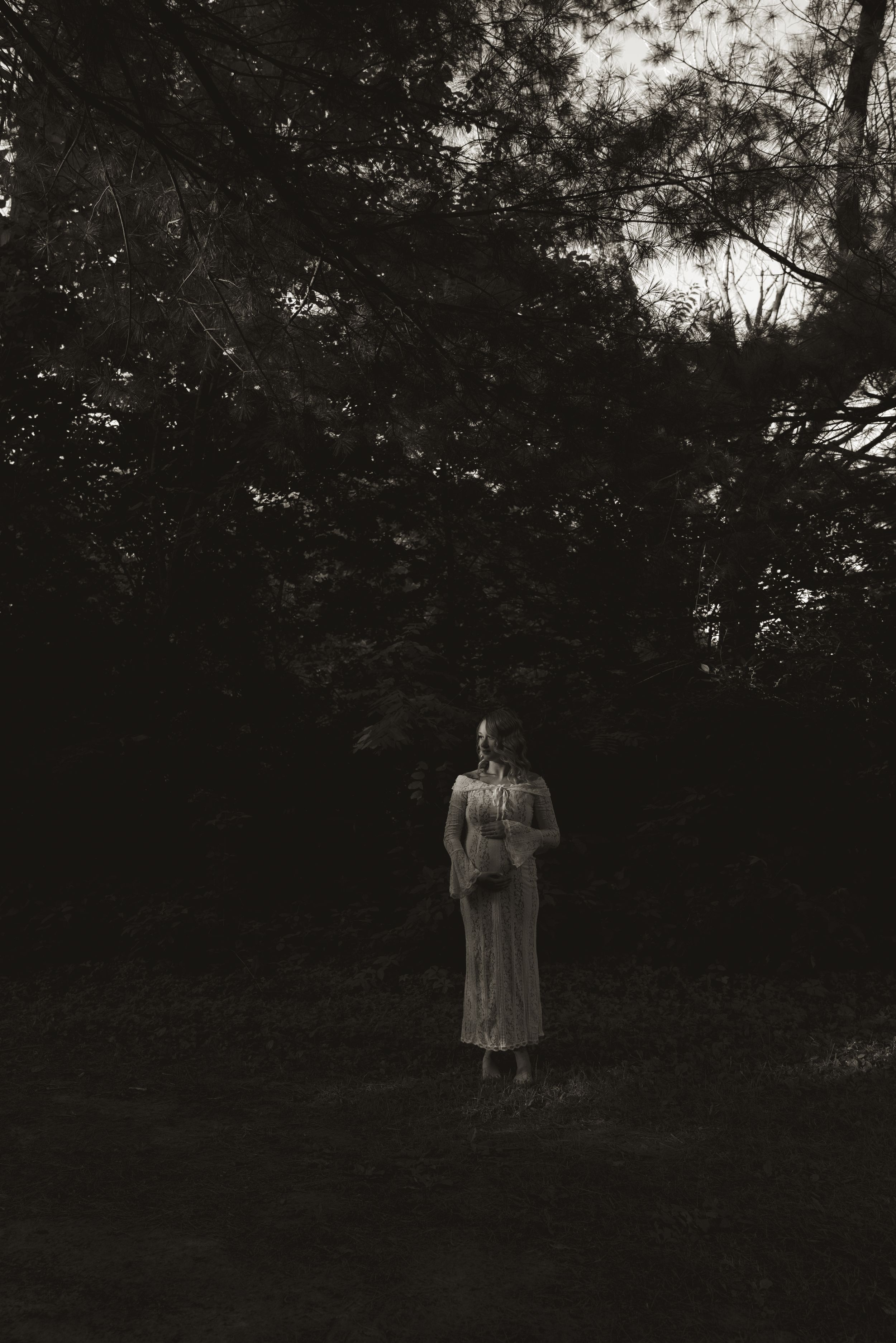 A silhouetted figure stands in a dark wooded area wearing a flowing dress.