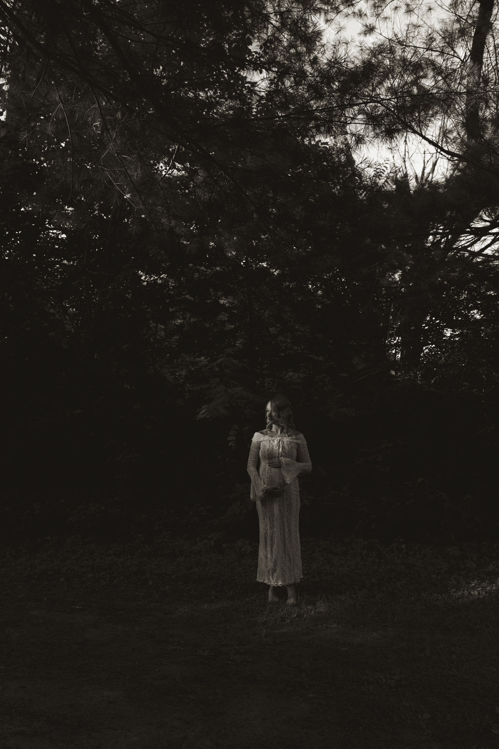 A silhouetted figure stands in a dark wooded area wearing a flowing dress.
