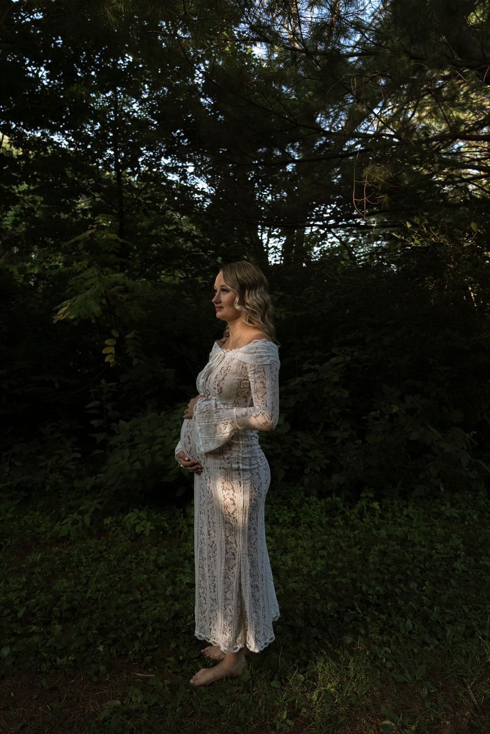 A person in a white lace dress stands in dappled sunlight among trees.