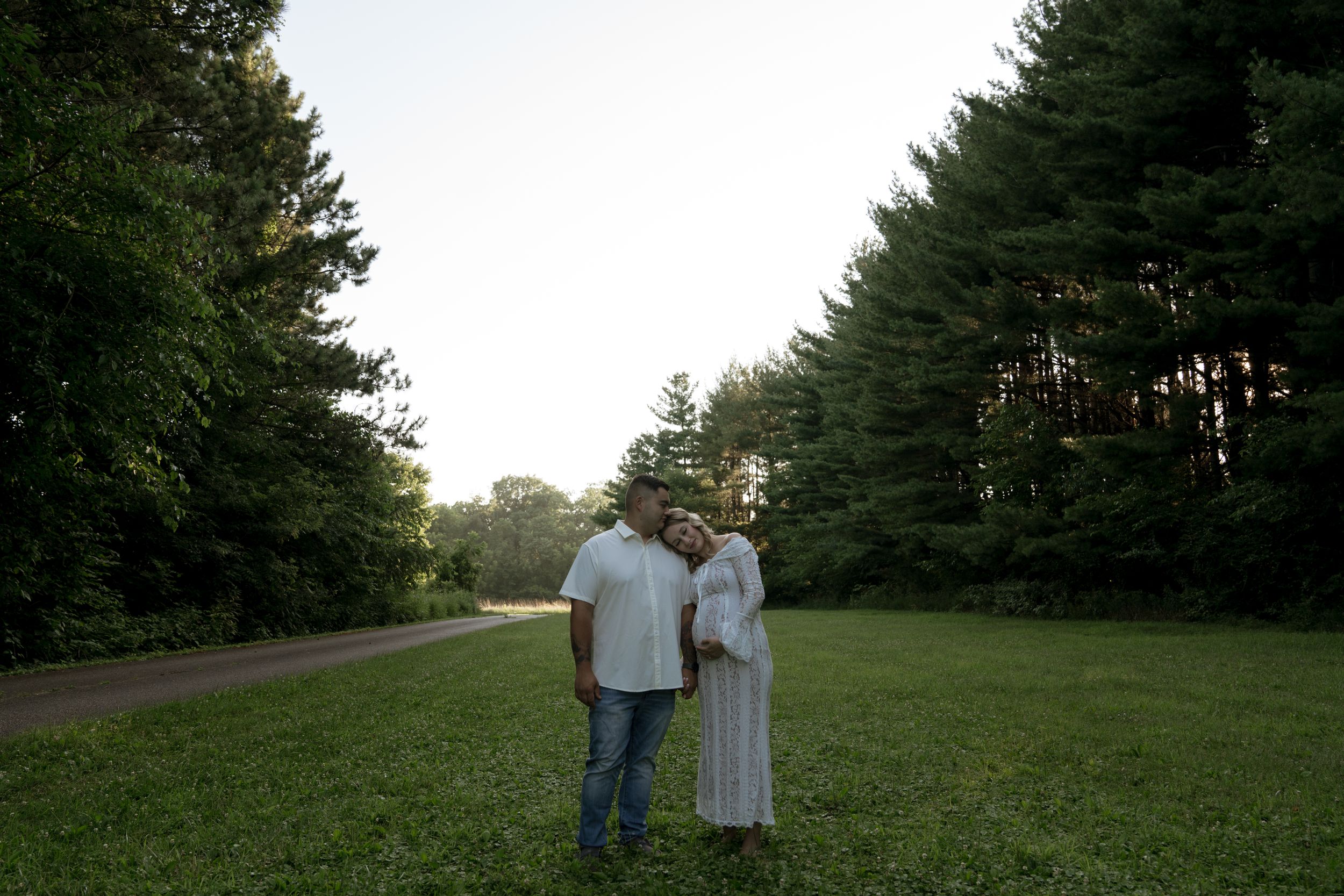 A couple stands together on a grassy hillside surrounded by pine trees.