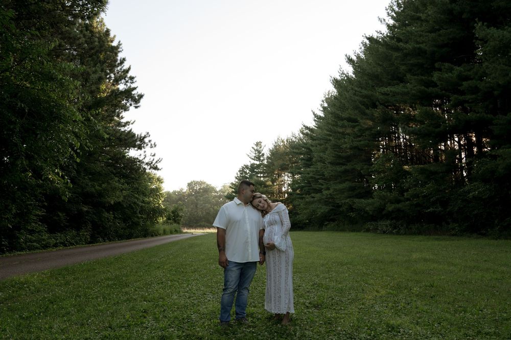 A couple stands together on a grassy hillside surrounded by pine trees.