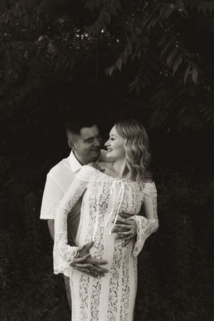 A romantic couple in white outfits share an intimate moment against a dark natural background.