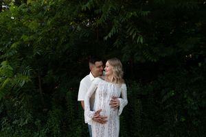 A couple embraces outdoors wearing coordinating white outfits among green foliage.