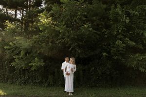 A couple in white clothing poses together in a wooded outdoor setting.