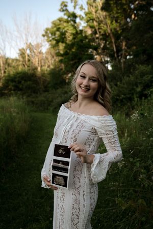 Woman in white lace dress holds up ultrasound photos while smiling outdoors.