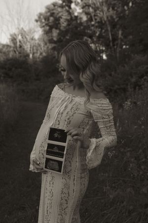 Woman in white lace dress shows ultrasound images during a pregnancy announcement photoshoot.