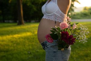 A pregnant woman in a white lace bralette holds a bouquet of pink and burgundy flowers at sunset in a grassy field.