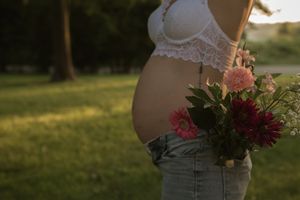 Silhouetted pregnant belly with floral bouquet against golden sunset light in outdoor setting.