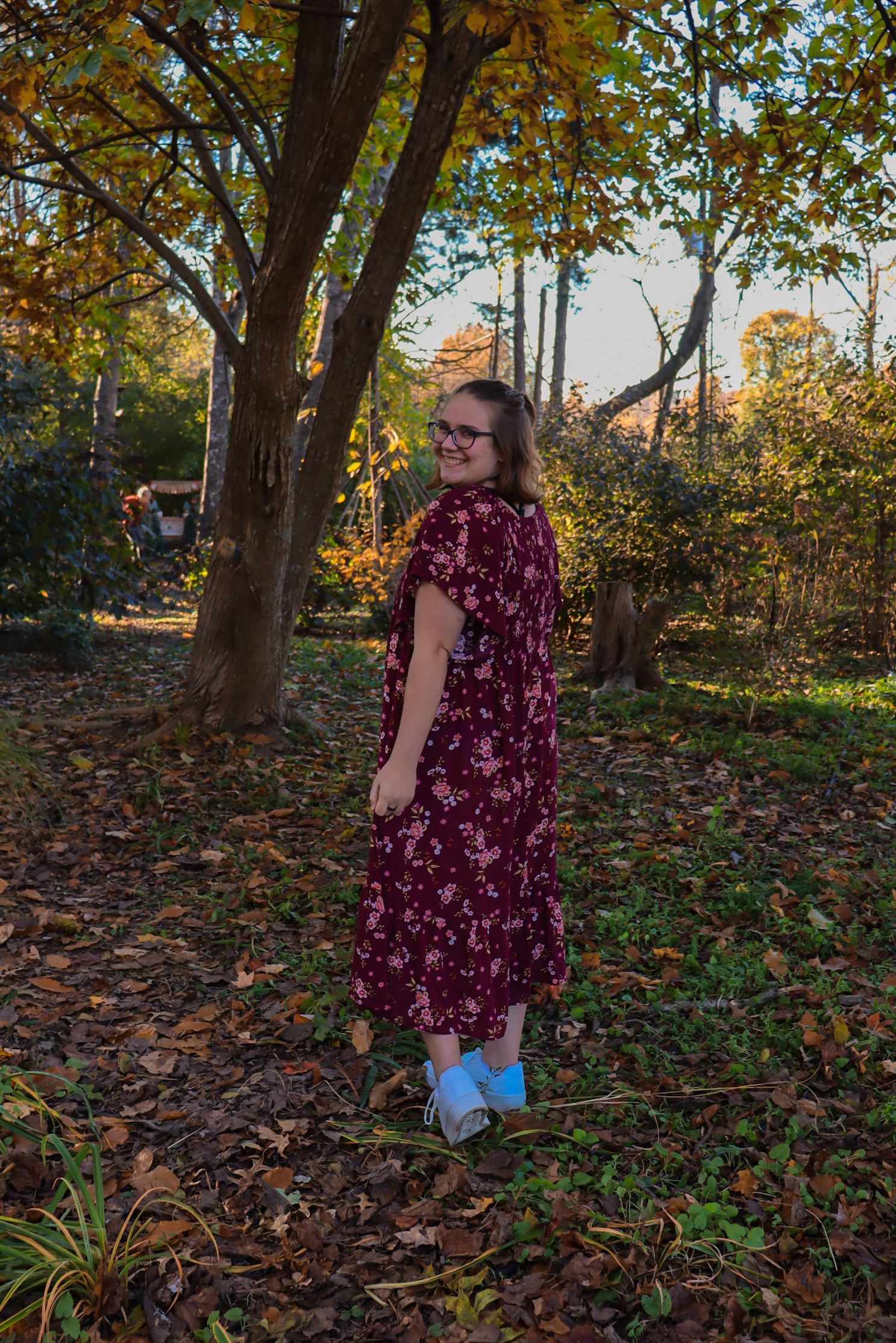 A figure in a burgundy floral dress and blue shoes stands in an autumn forest setting with fallen leaves on the ground.
