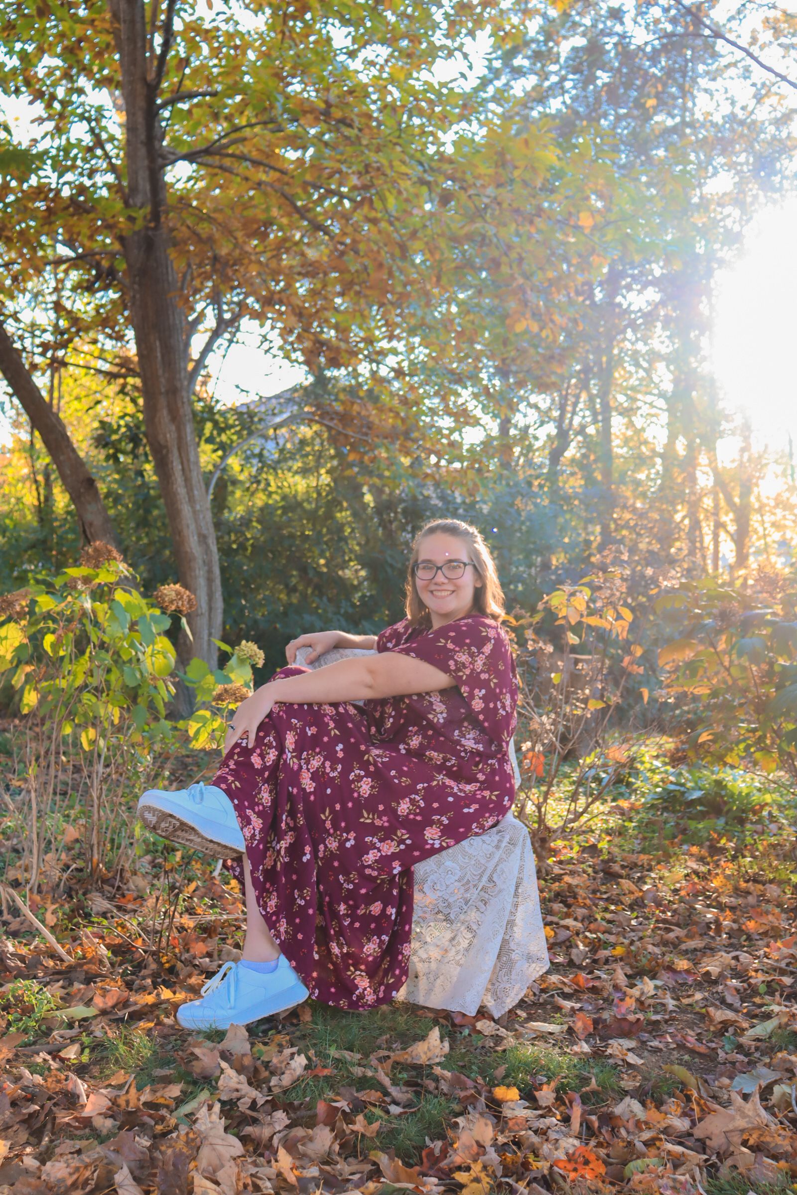 Woman in burgundy floral dress sits on blanket among fallen autumn leaves during golden hour sunset in wooded setting.