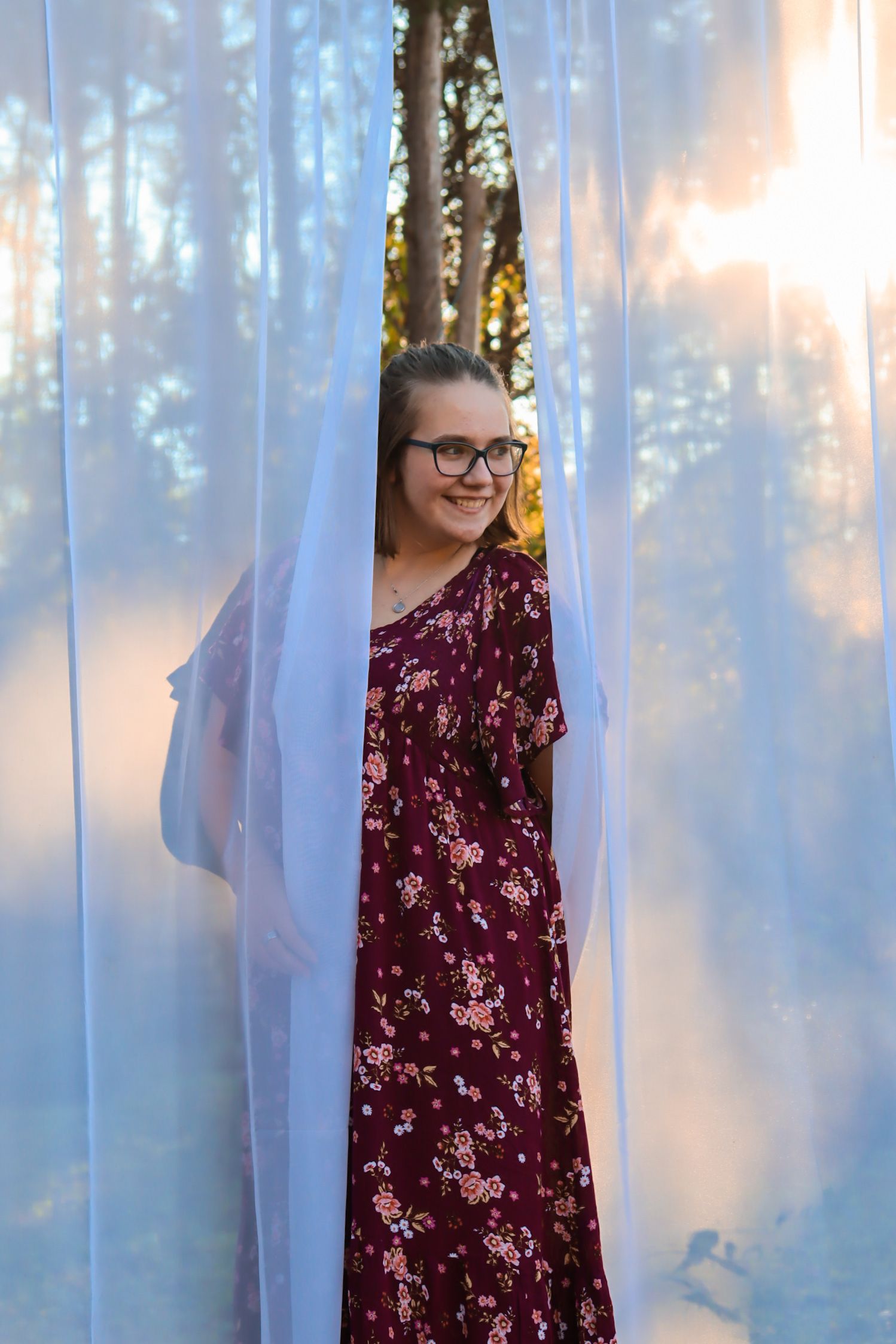A person wearing glasses and a burgundy floral dress poses between sheer white curtains in an outdoor setting.