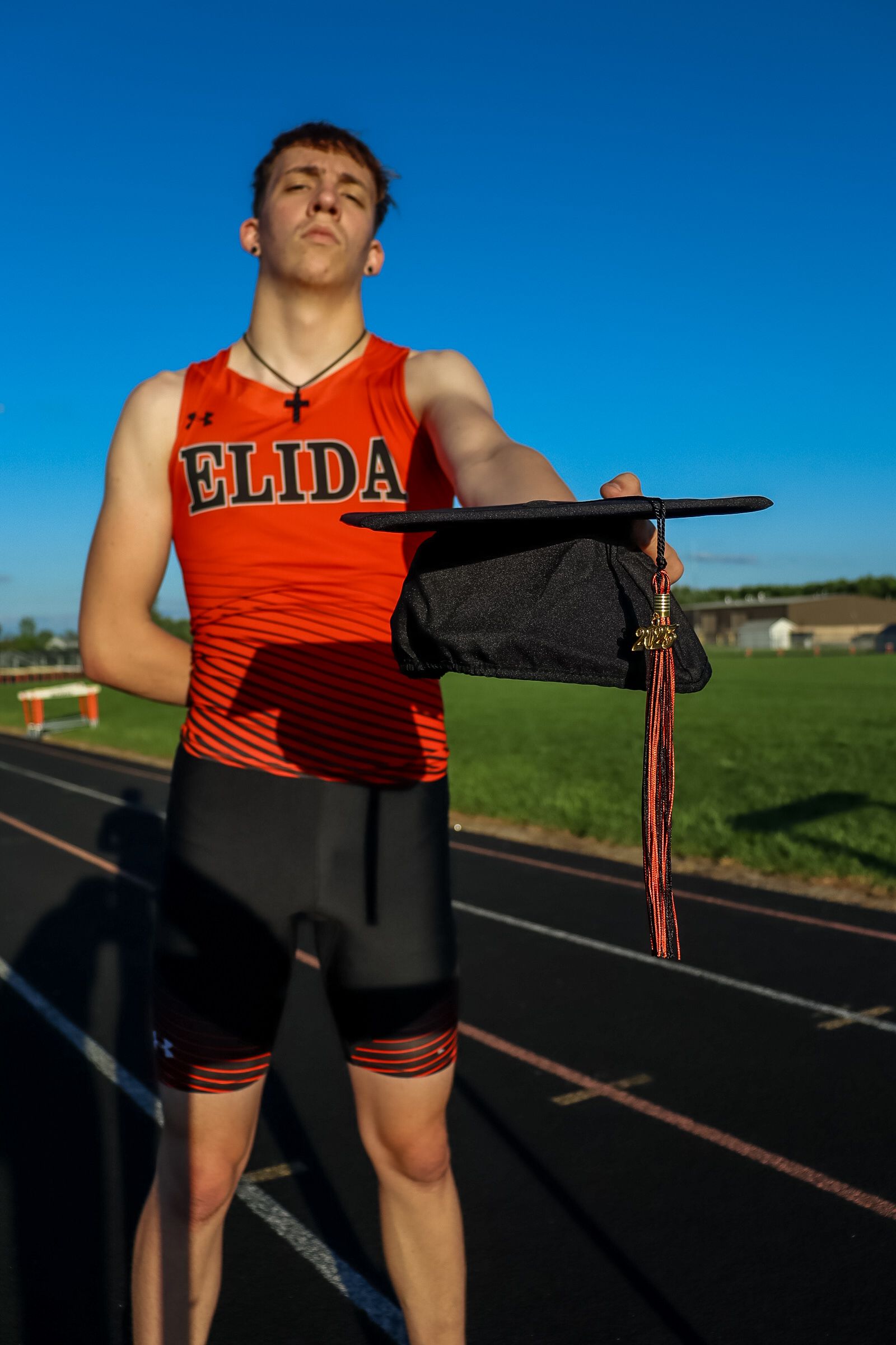 Athlete in an orange and black uniform stands beneath the graduation cap raised against a bright blue sky background.