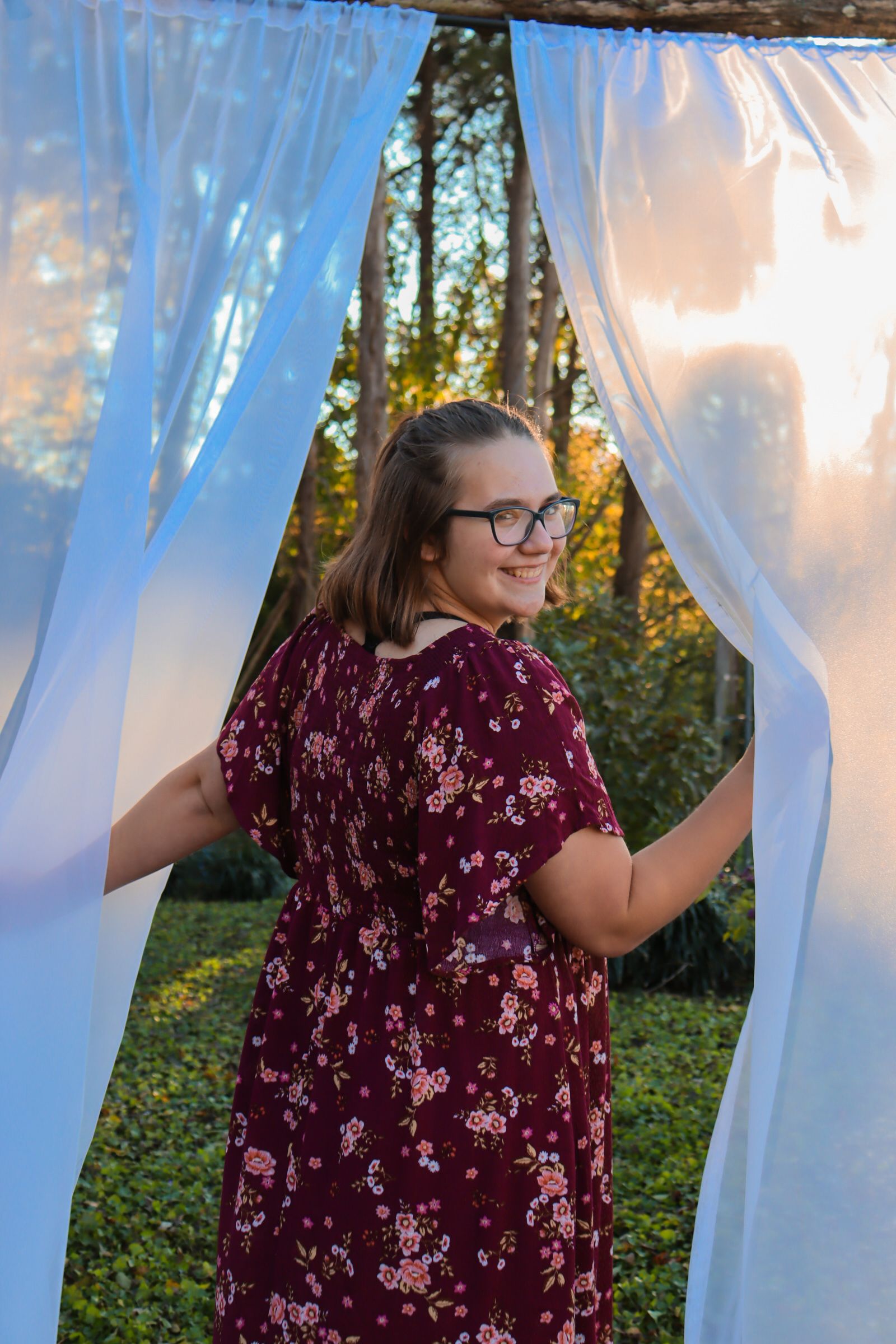 A person in a burgundy floral dress stands between flowing white and blue curtains in an outdoor garden setting at sunset.