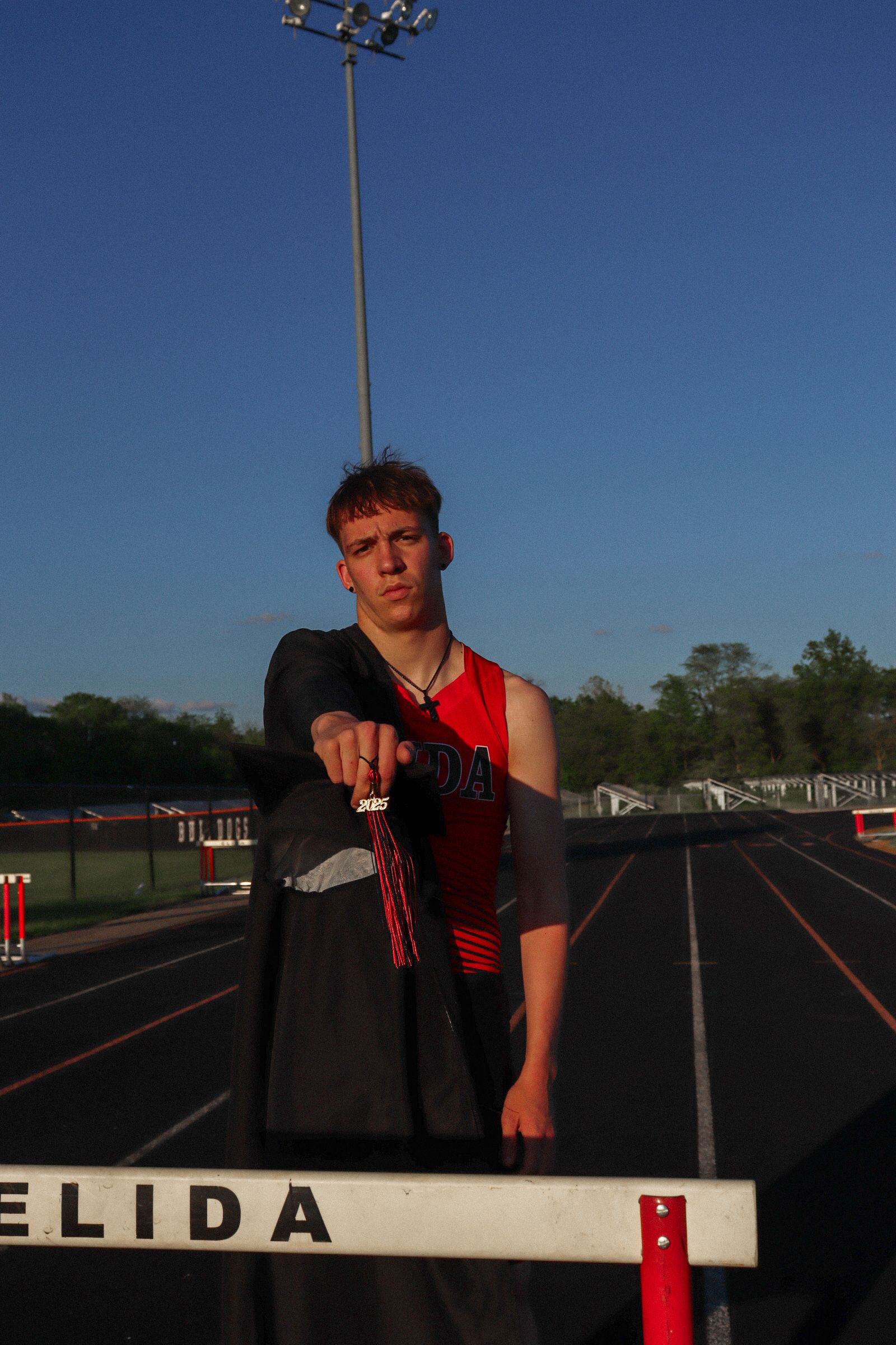 An athlete stands beside a track hurdle at dusk with stadium lights and a blue sky in the background.