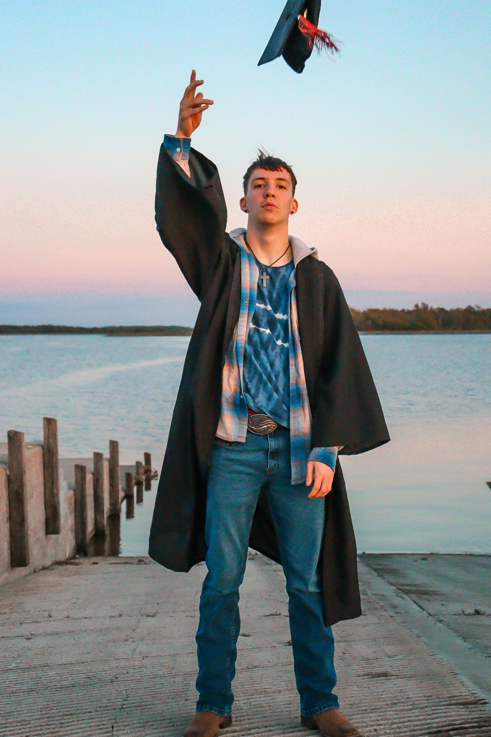 A graduating student in cap and gown poses joyfully on a wooden dock by a tranquil lake at sunset.