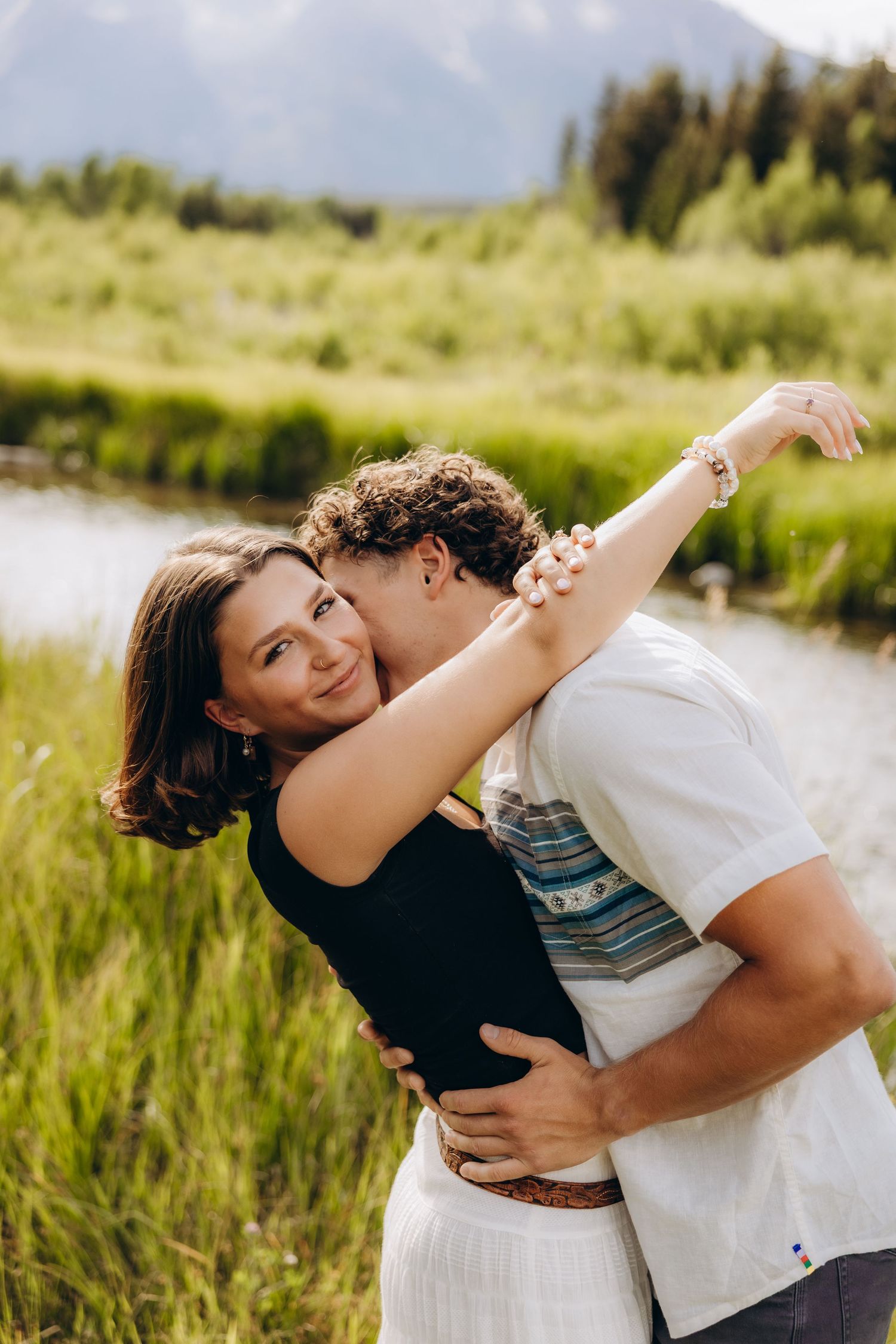 A romantic couple embraces in a sun-lit meadow near a lake with mountains and forest in the background.