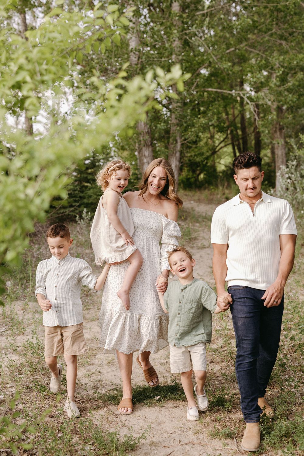 Family wearing coordinated neutral outfits walks together through a scenic wooded path during an outdoor photo session.
