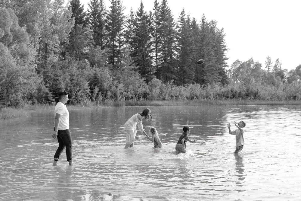 Vintage black and white photograph of people wading and splashing in a river with evergreen trees along the shoreline.