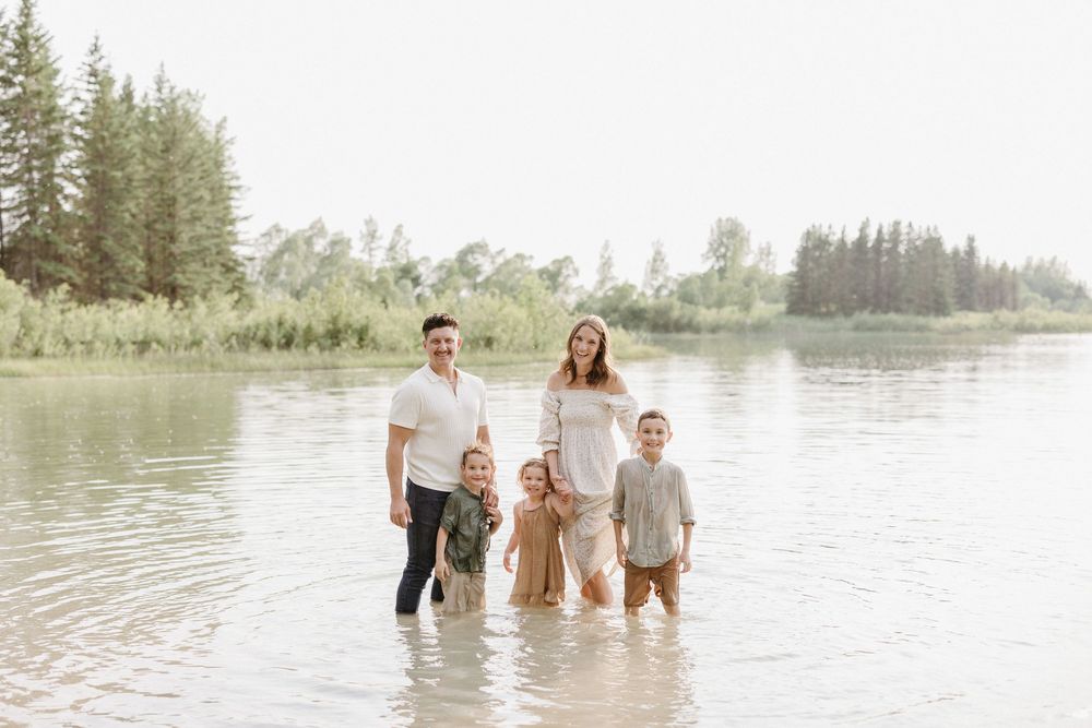 Family wading in calm lake water with evergreen trees and lush landscape in background.