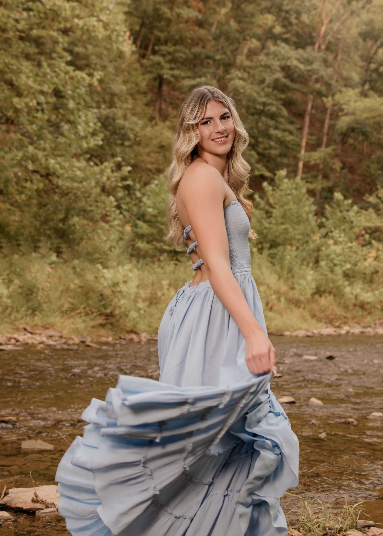 Figure in flowing blue dress poses by river with dramatic fabric movement.