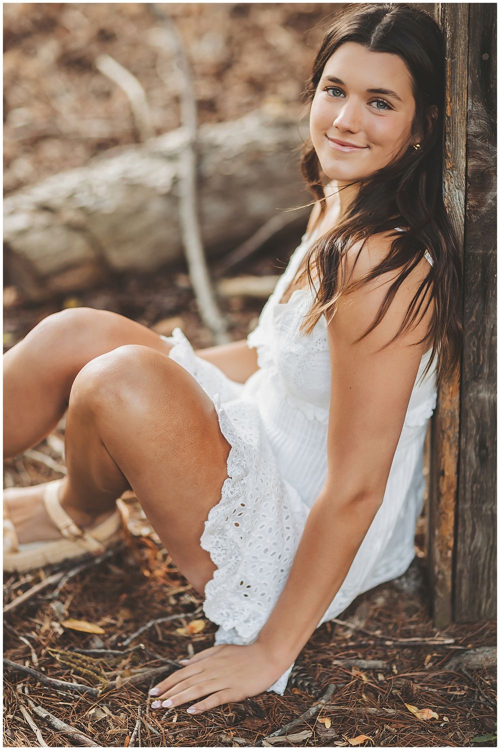 Candid poses in white eyelet dress against wooden post outdoors.