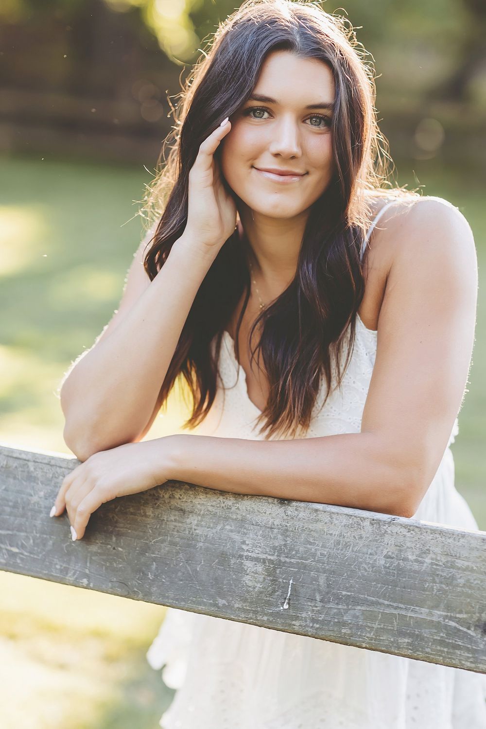 A woman in white leaning on a wooden fence in golden sunset lighting.