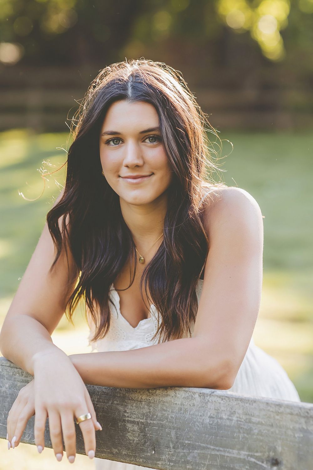 Portrait of a woman resting arms on a wooden fence with sun-dappled background.