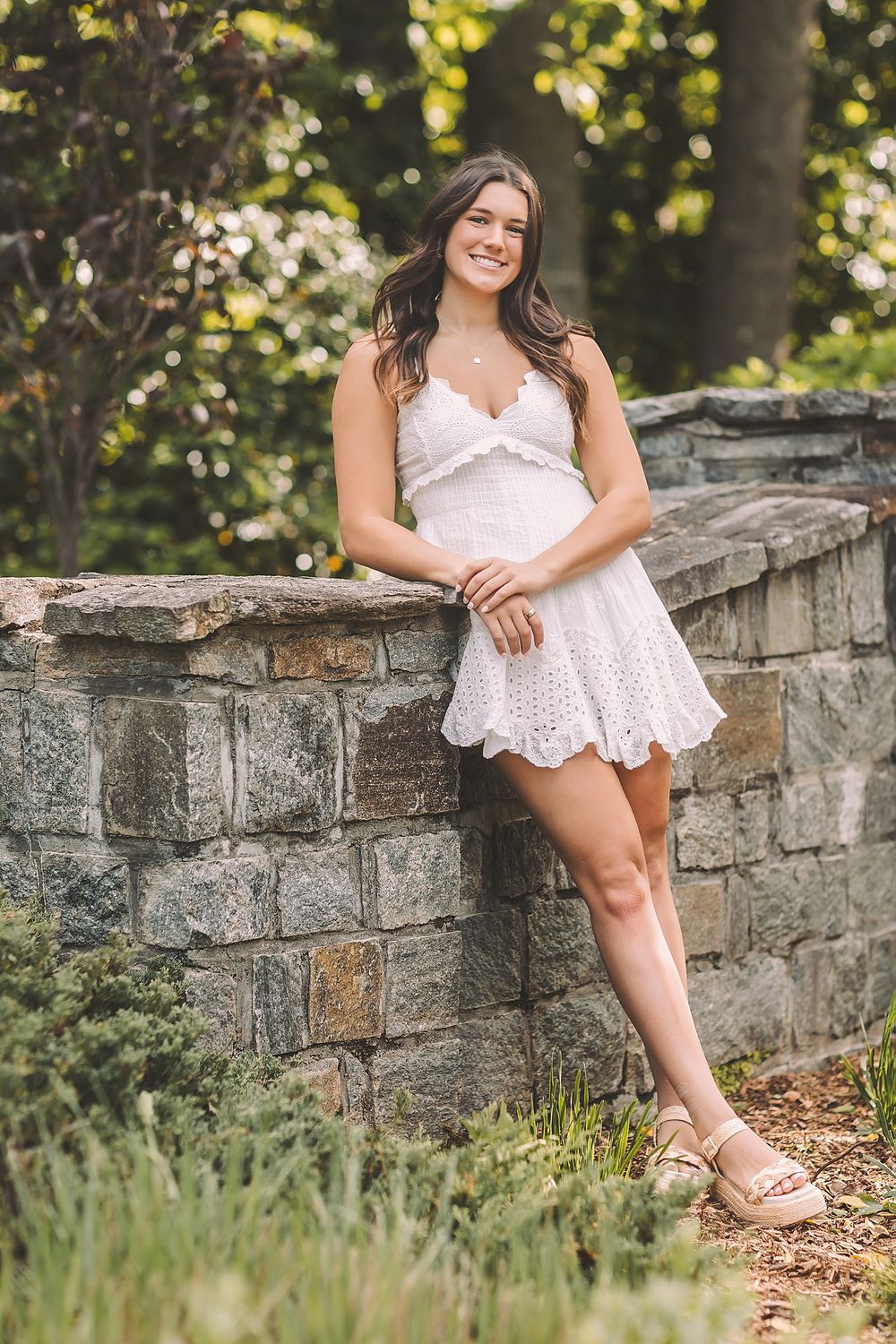 A summery portrait session features a white eyelet dress and stone wall backdrop in a garden setting.
