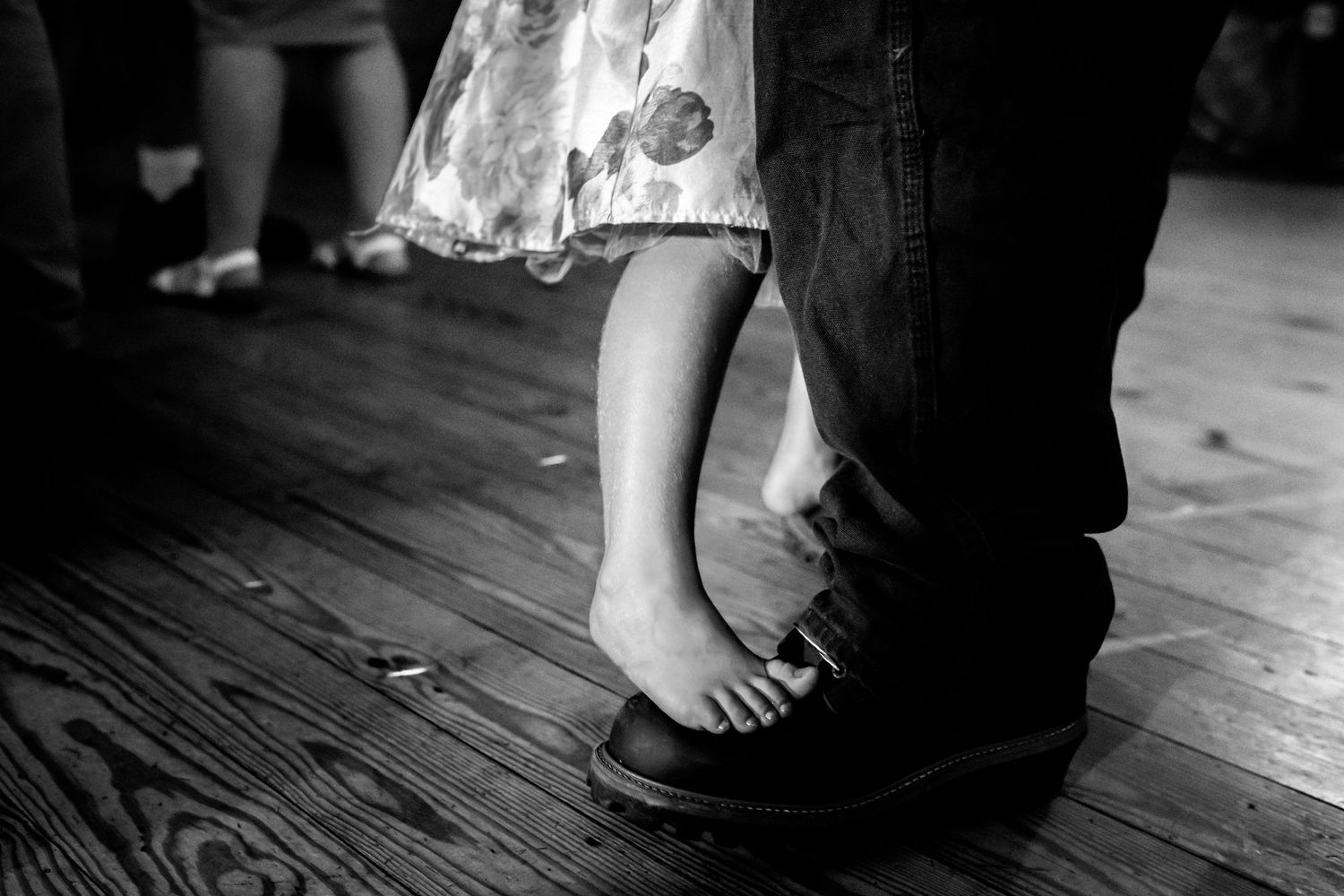 Close-up details of bare feet and formal attire on a wooden floor at a wedding.
