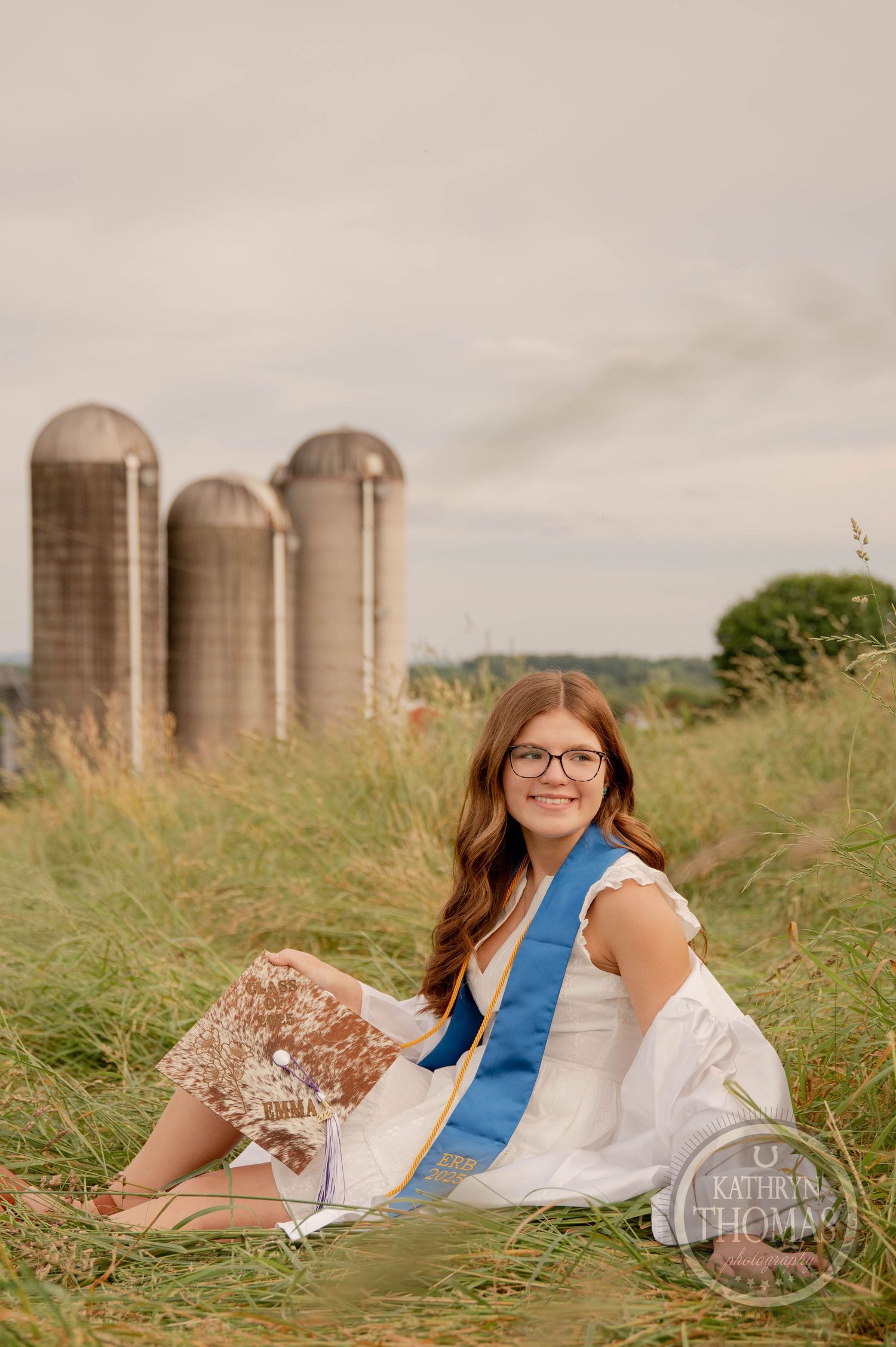 A white dress and blue sash complement the rustic scene with tall farm silos and grass in the background.