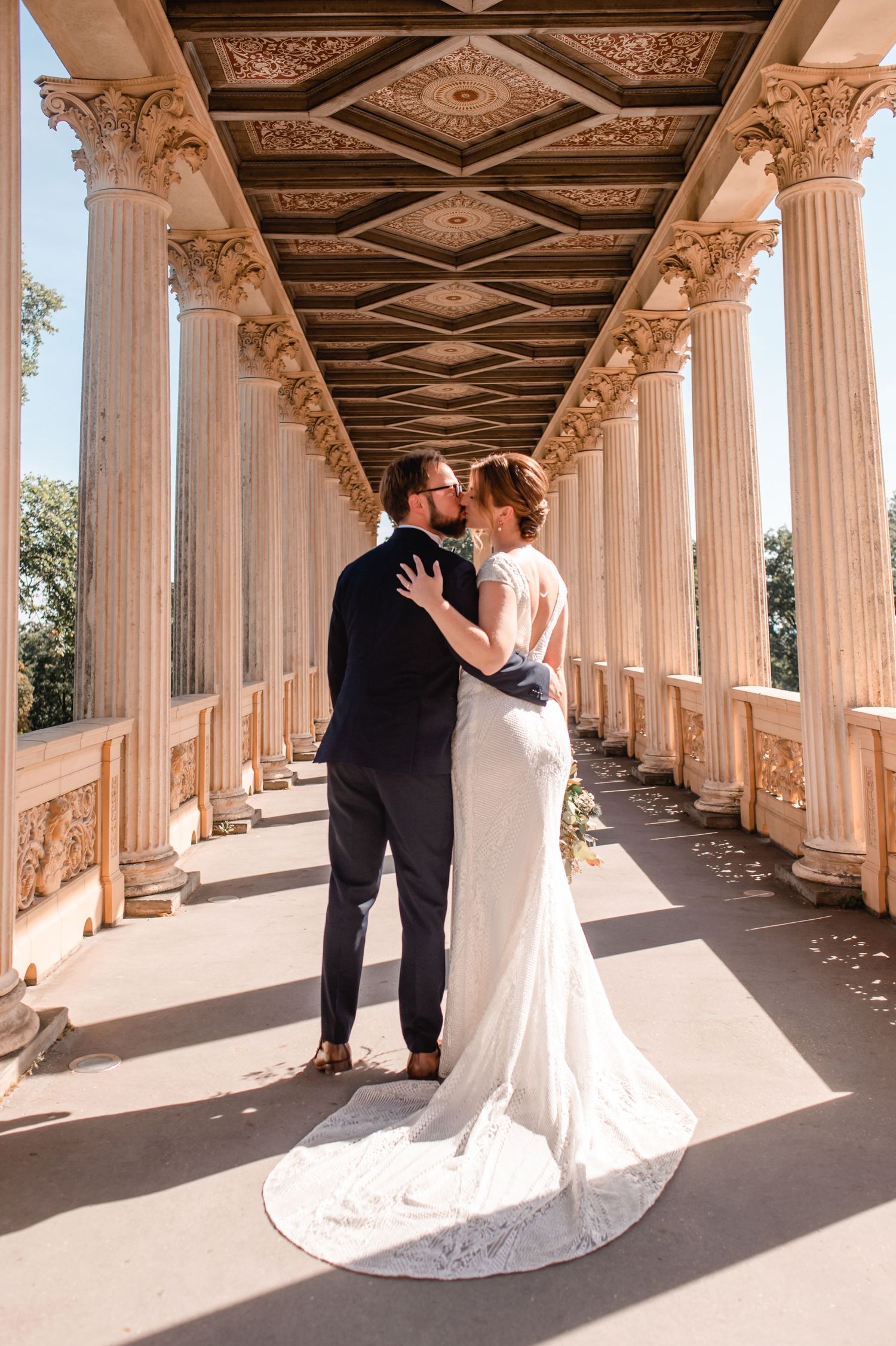 A couple in wedding attire poses under an ornate columned portico with a sweeping train and classic architecture.