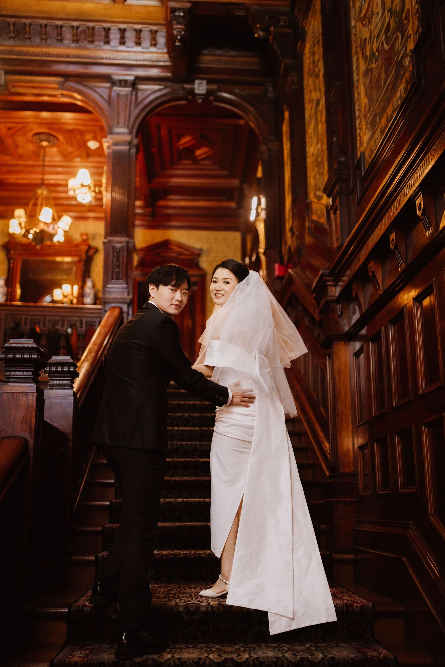 Wedding couple poses on ornate wooden staircase in historic building with vintage lighting.