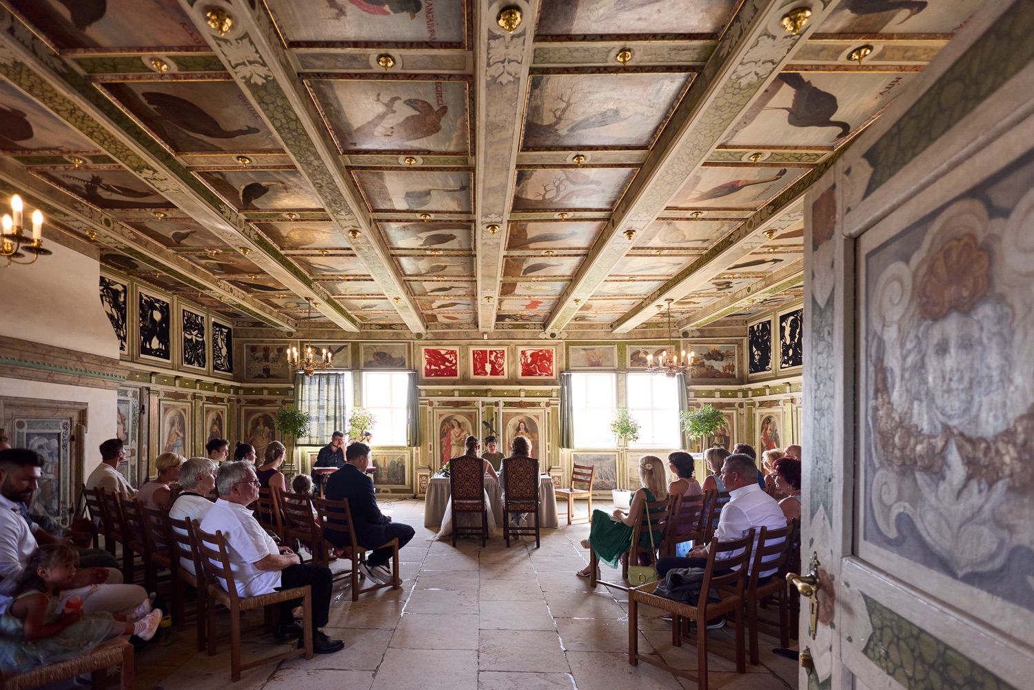 A historical ceremony taking place in an ornate room with wooden coffered ceiling and side murals as attendees fill the pews.