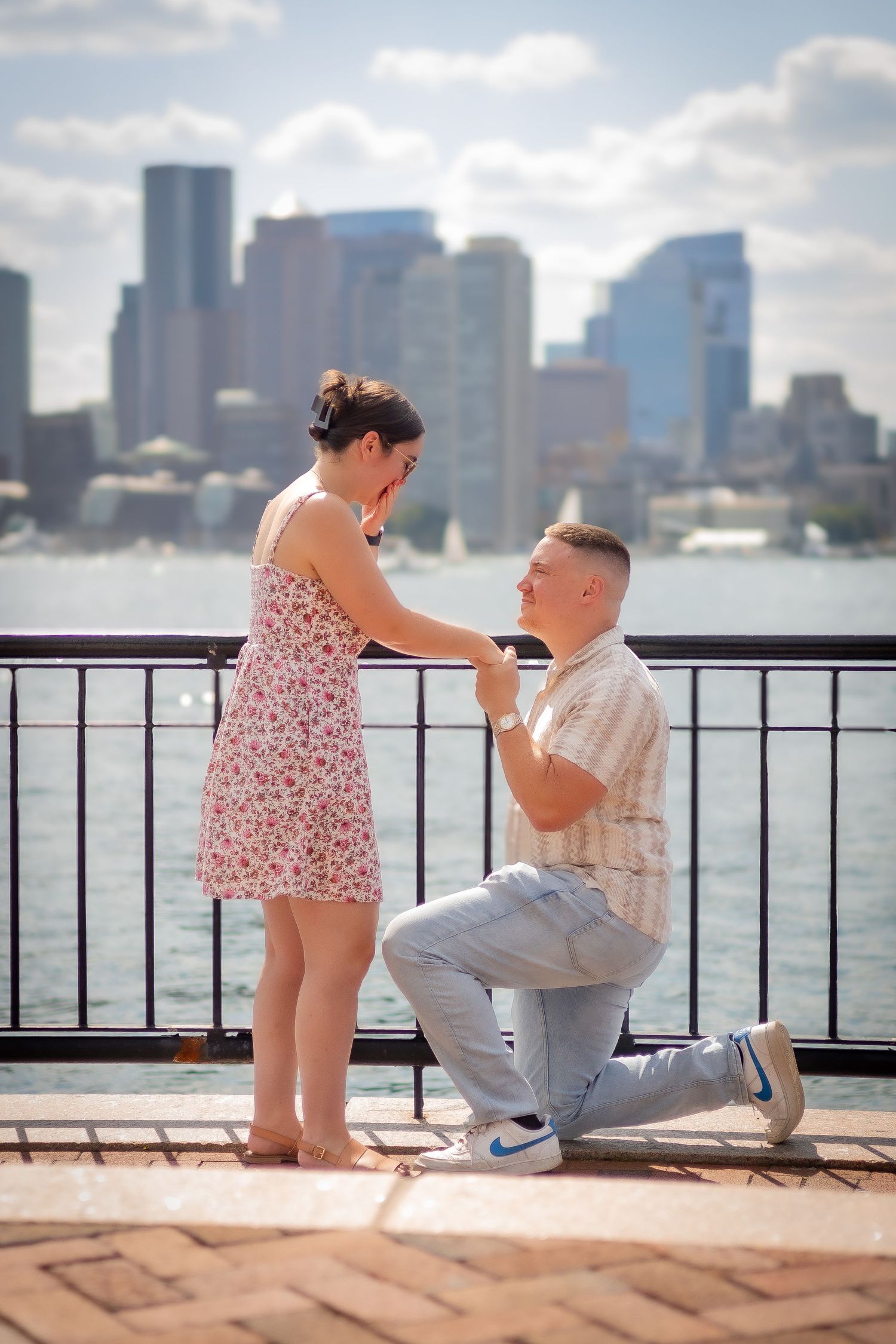 A romantic marriage proposal scene unfolds on a waterfront deck overlooking a cityscape on a sunny day.