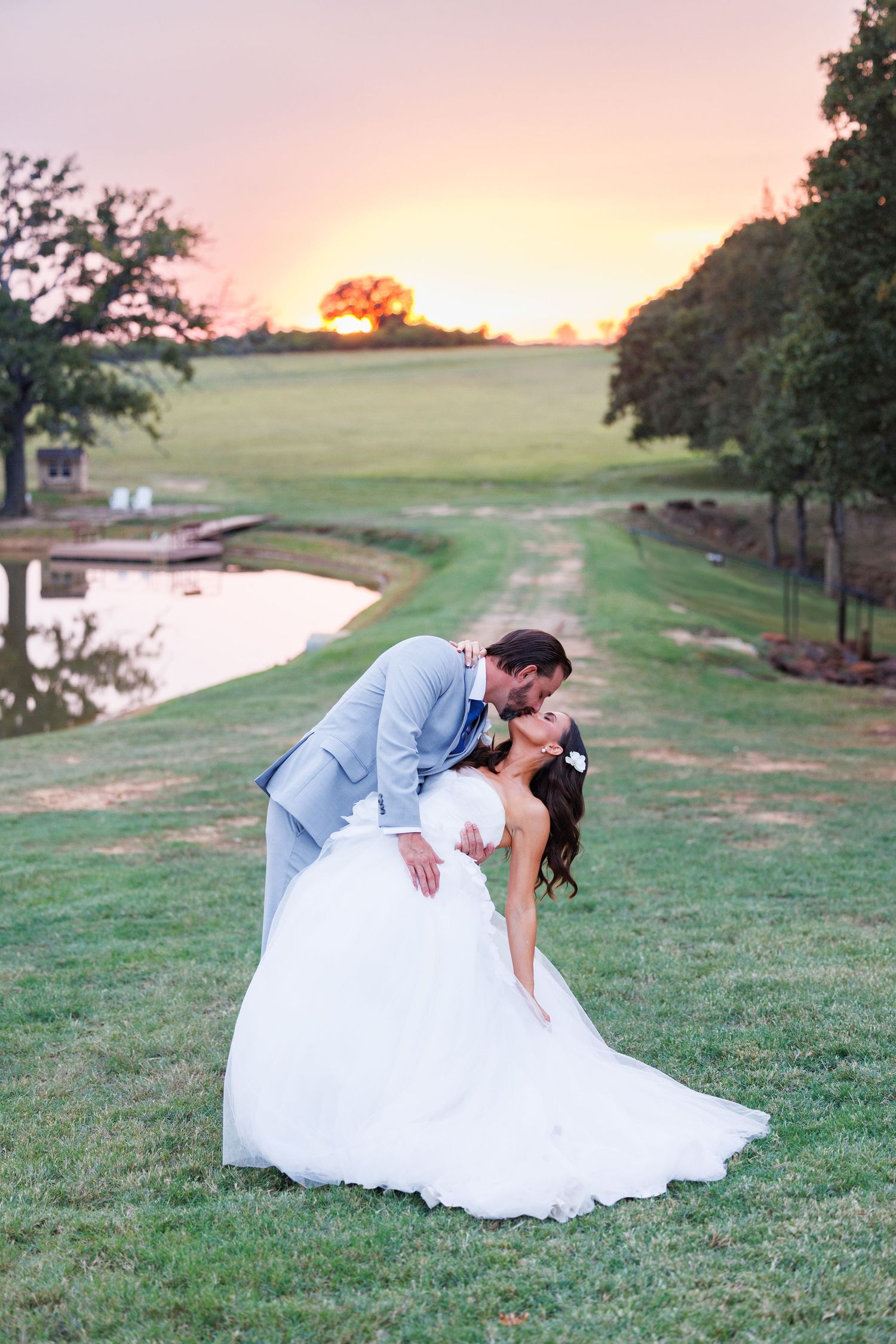A bride and groom in wedding attire kiss against a sunset backdrop at Bella Cavalli with orange sky and trees in background.