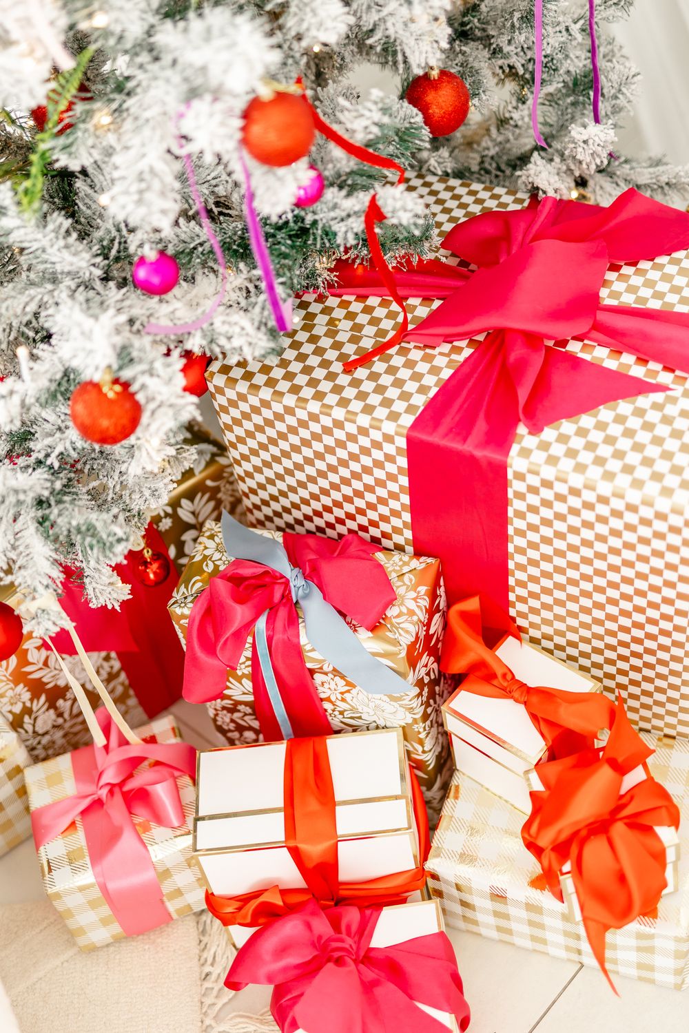 Beautifully wrapped Christmas presents with red bows sit beneath a snow-dusted evergreen tree with twinkling lights.