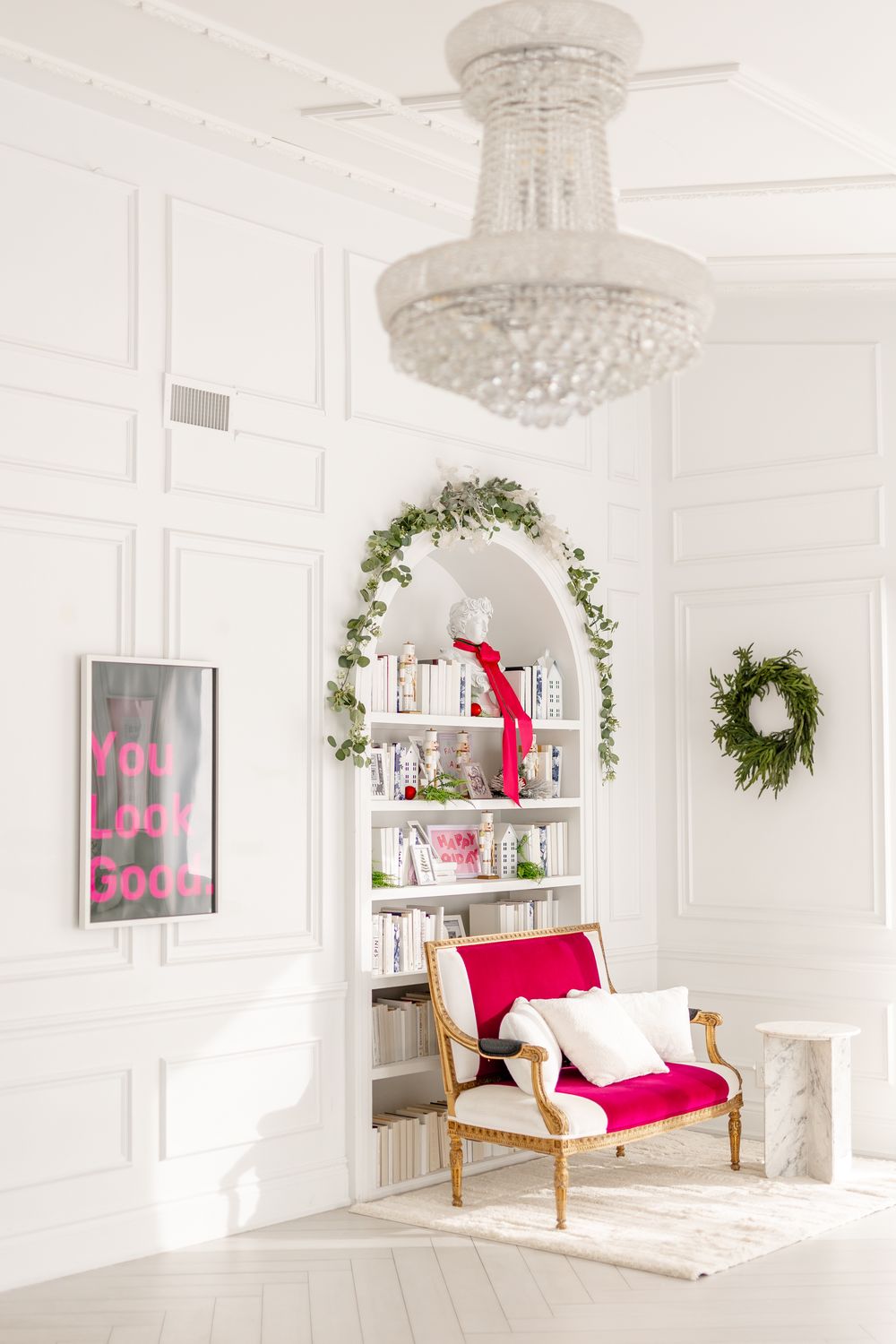 Bright white room with crystal chandelier, red accent chair, decorative greenery arch over bookshelf, and holiday wreath decor.