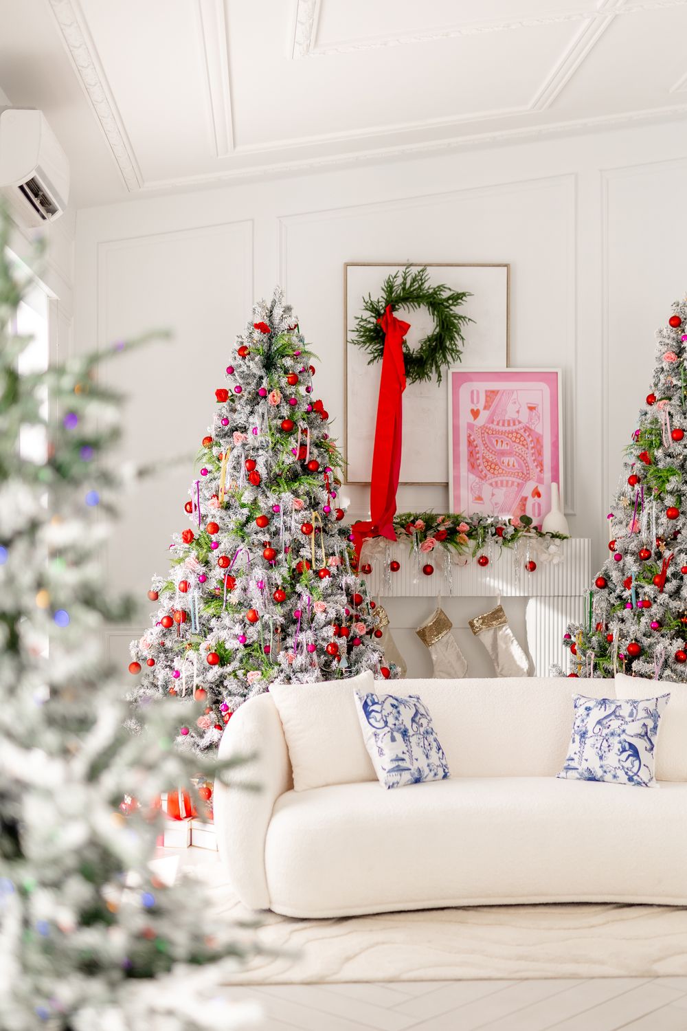 Multiple decorated Christmas trees flank a cream colored sofa with blue and white pillows in a bright white room.