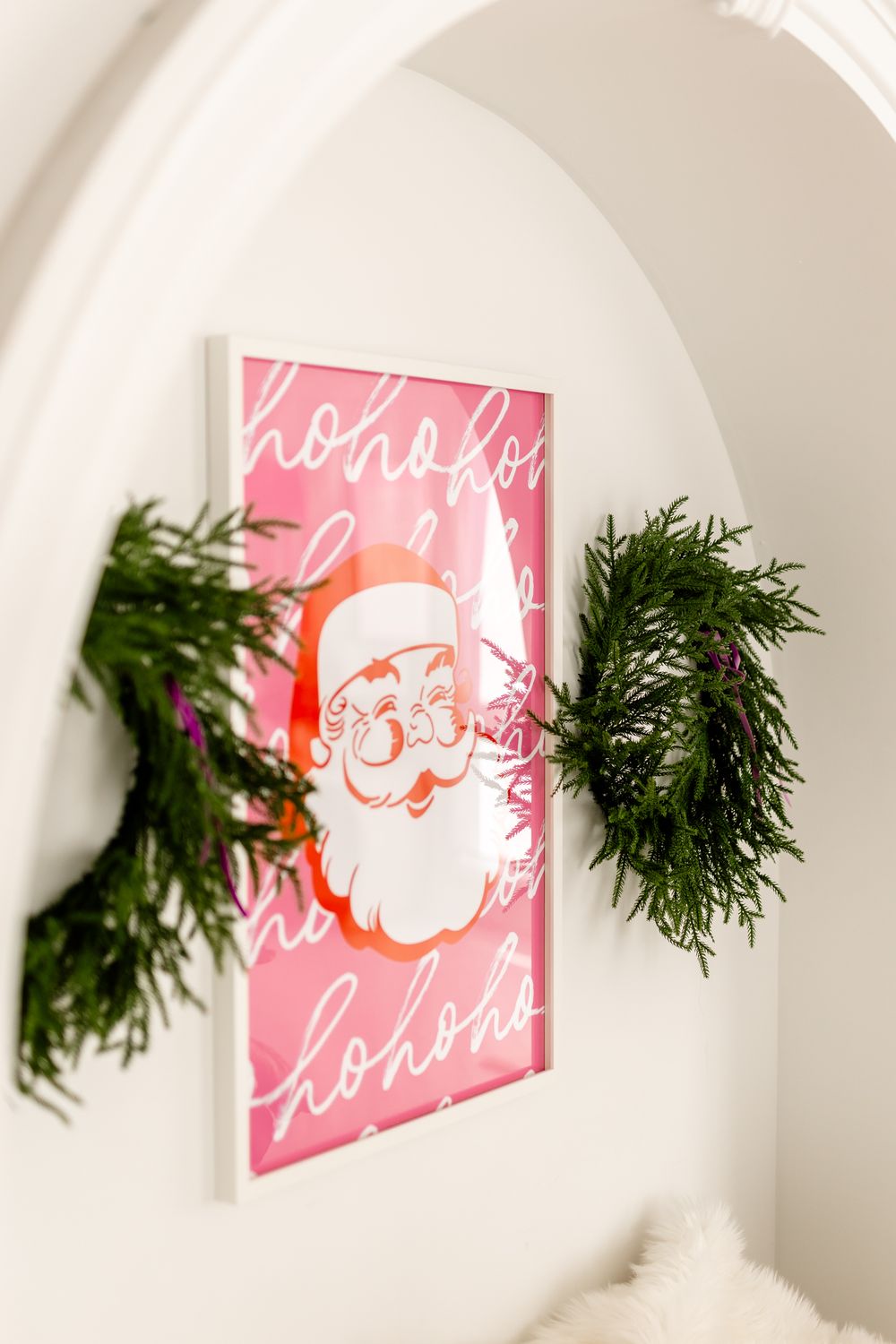 Pink holiday wall art featuring Santa Claus design with green evergreen branches decorating an arched white wall.