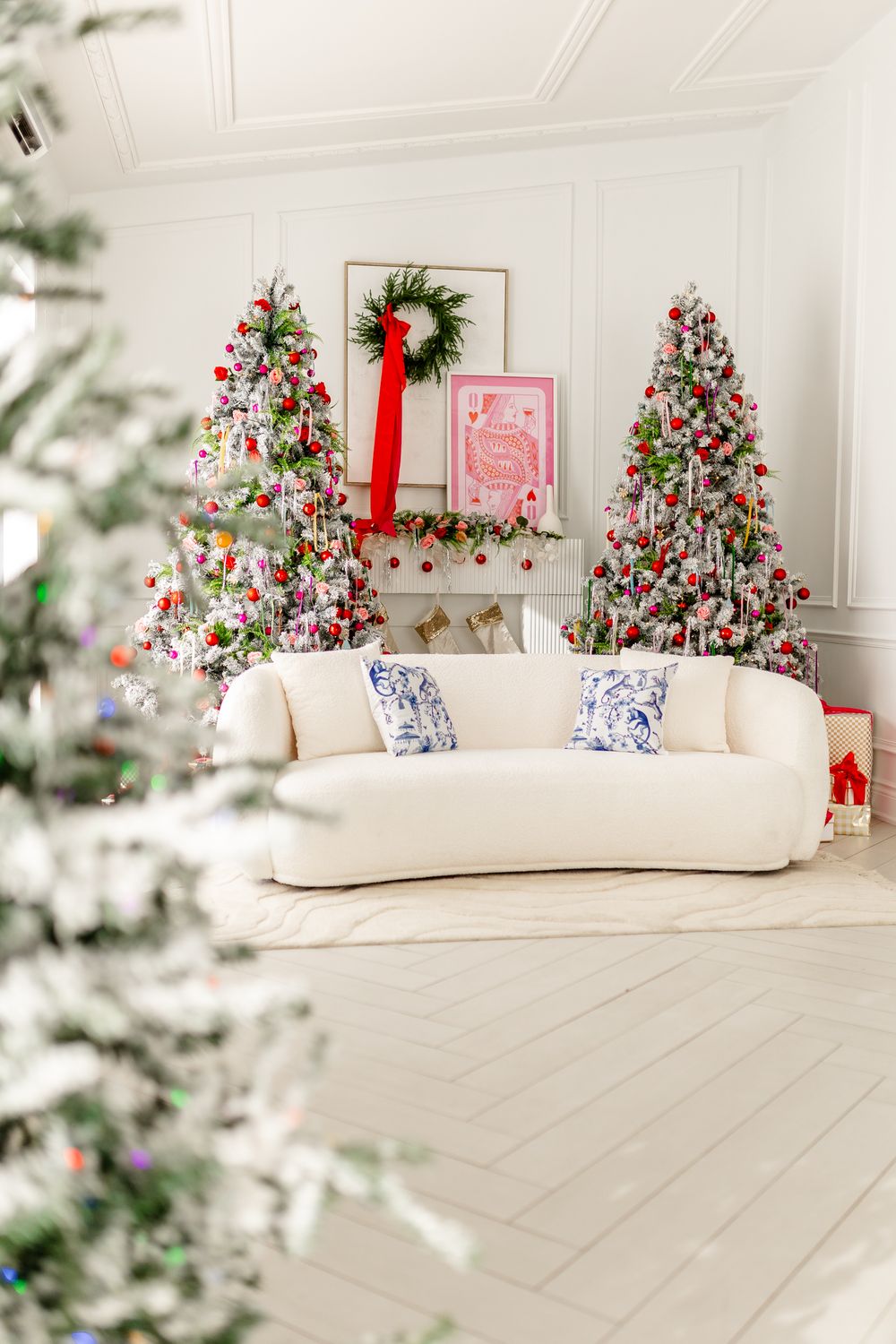Elegant white living room with three Christmas trees decorated in red and white, featuring a curved cream sofa and festive wreath.