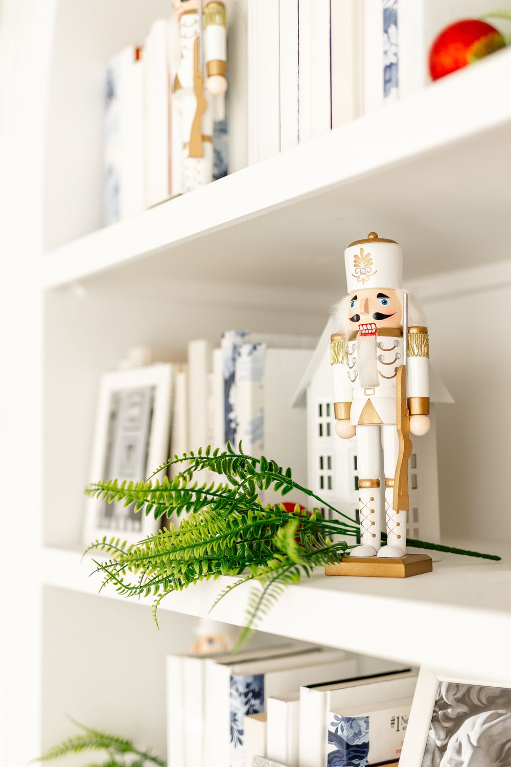 White decorative nutcracker figure displayed on modern white shelving with fern greenery and books as holiday decor.
