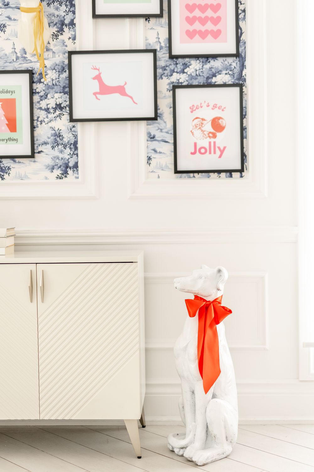 A white wall with framed pink artwork and a white ceramic dog statue wearing a red bow.