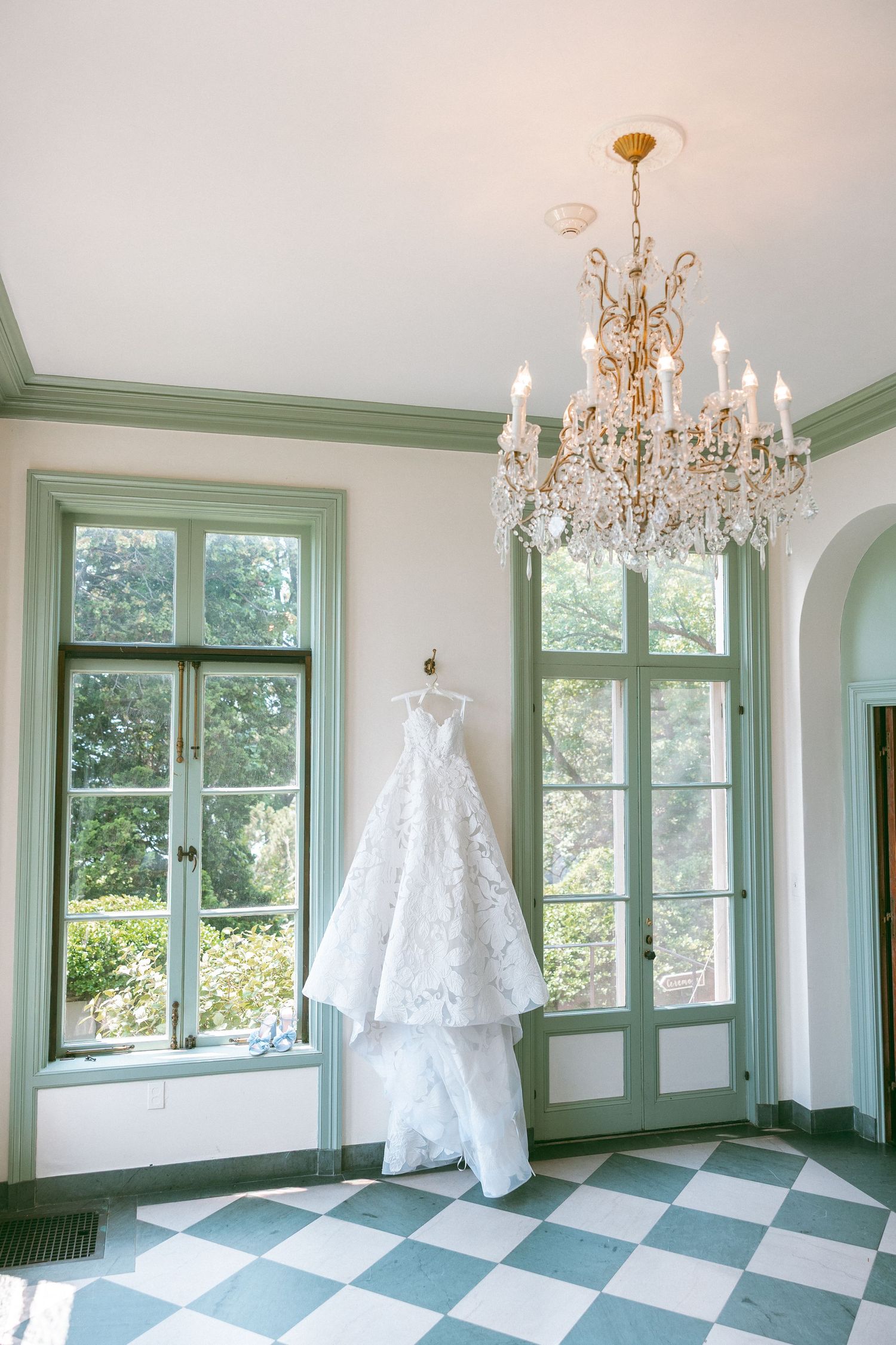 Elegant room with crystal chandelier and white wedding dress displayed by window.