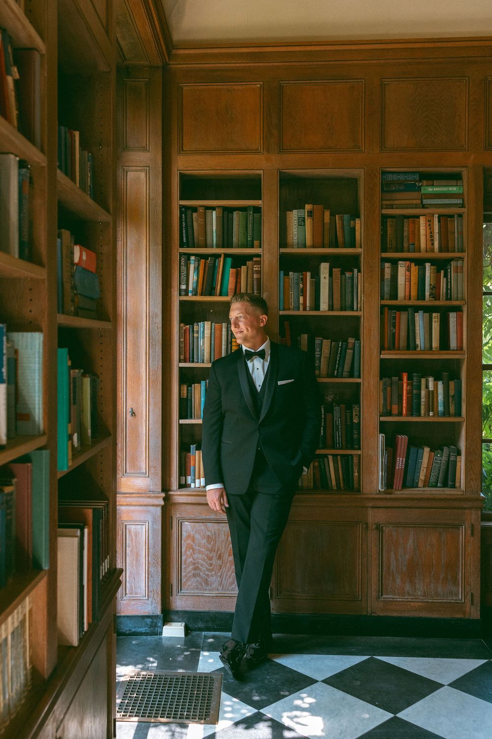 A groom poses progressively in a wood-paneled library with built-in bookshelves.