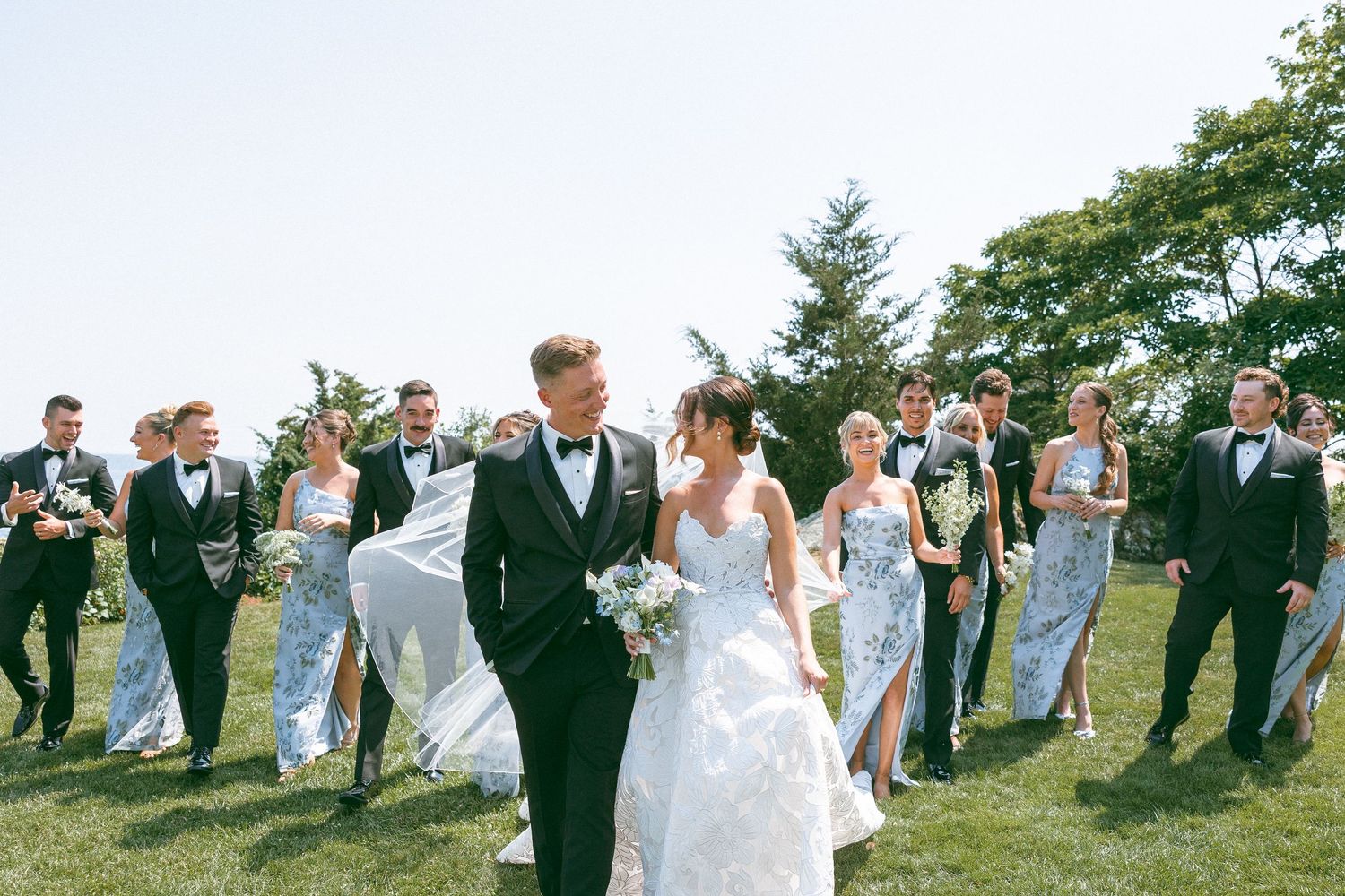 Wedding party runs and poses playfully on a grassy hillside overlooking the ocean.