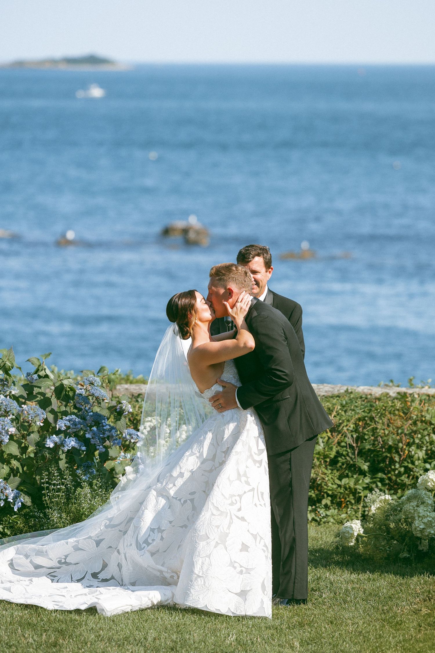 A romantic wedding couple shares a kiss on a grassy oceanfront lawn with blue waters in the background.