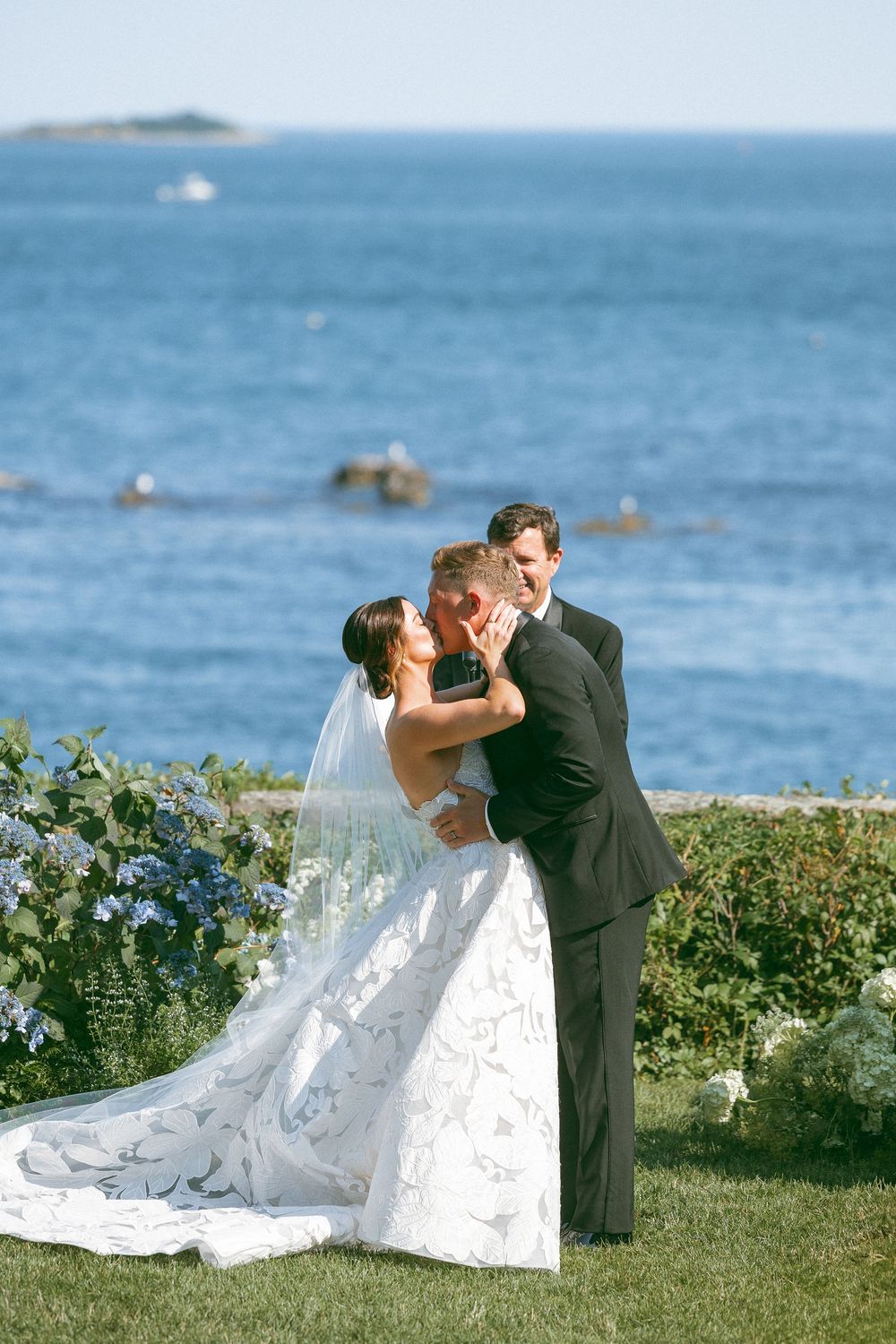 A romantic wedding couple shares a kiss on a grassy oceanfront lawn with blue waters in the background.