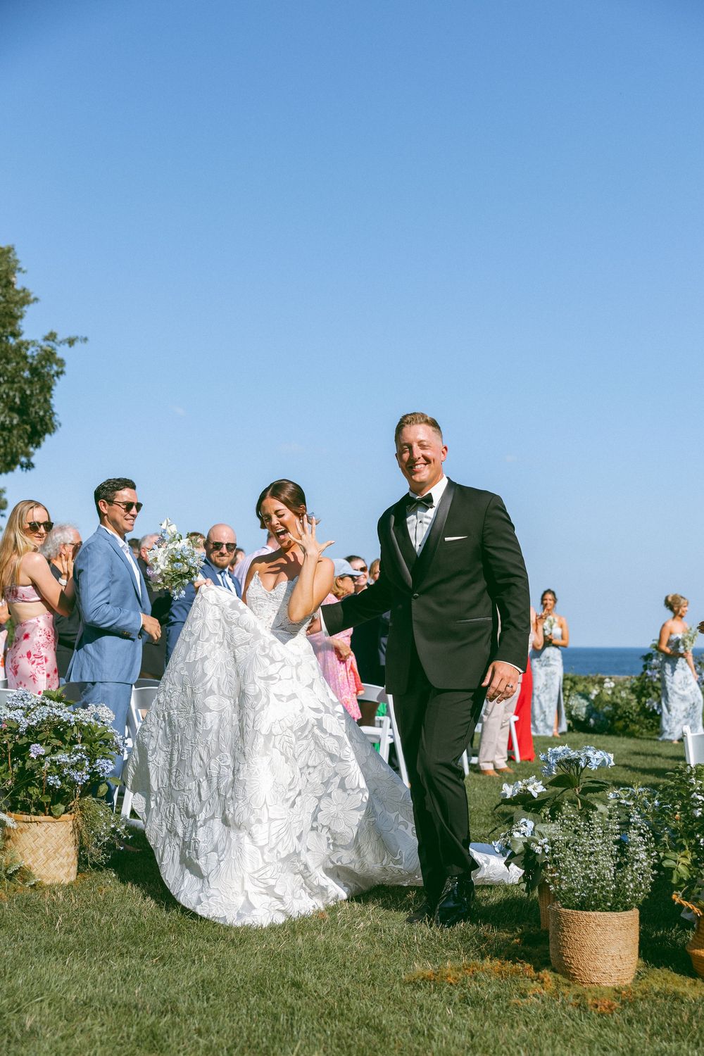 Wedding guests gather on a seaside lawn as a couple walks through in formal attire.
